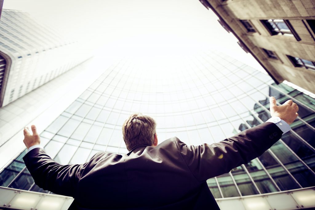 A businessman outside with arms out stretched, looking upward at office building